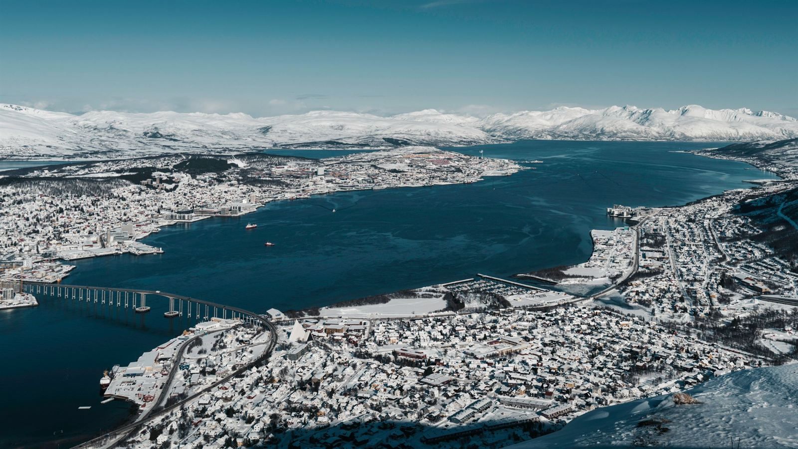 Snow-covered Tromsø on its island in a blue fjord, with the long harbor bridge and white Arctic mountains on the horizon