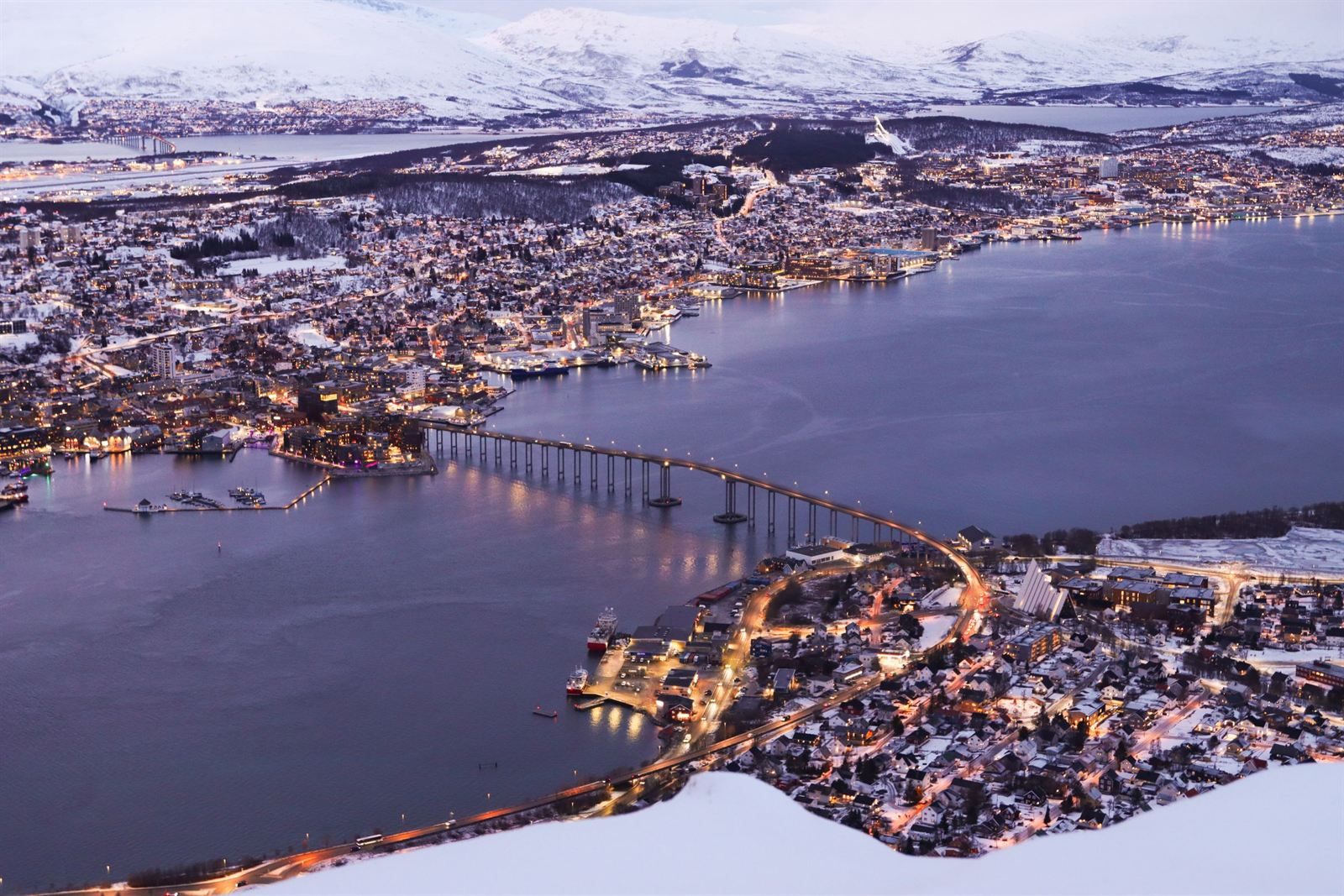 Tromsø at twilight from the mountainside, with the city lights, the arched bridge across the sound and snowy peaks beyond